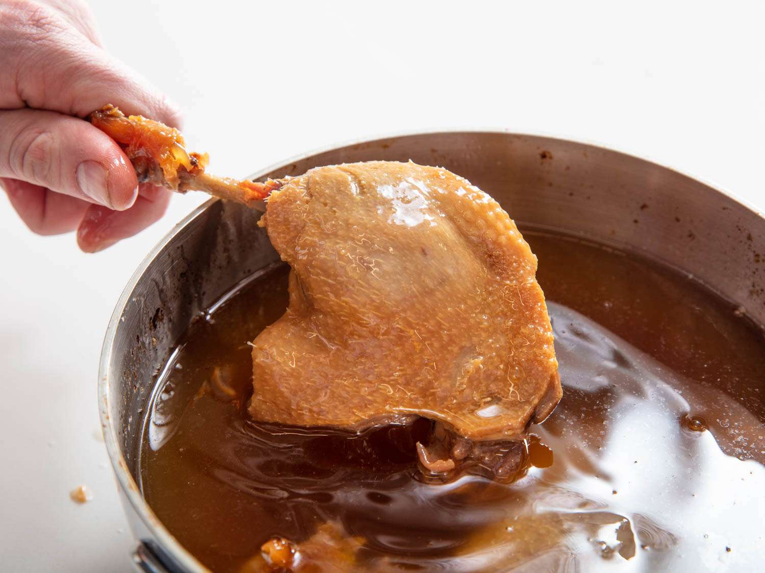 A leg of koji duck confit is held above the surface of the duck fat it was cooked in.