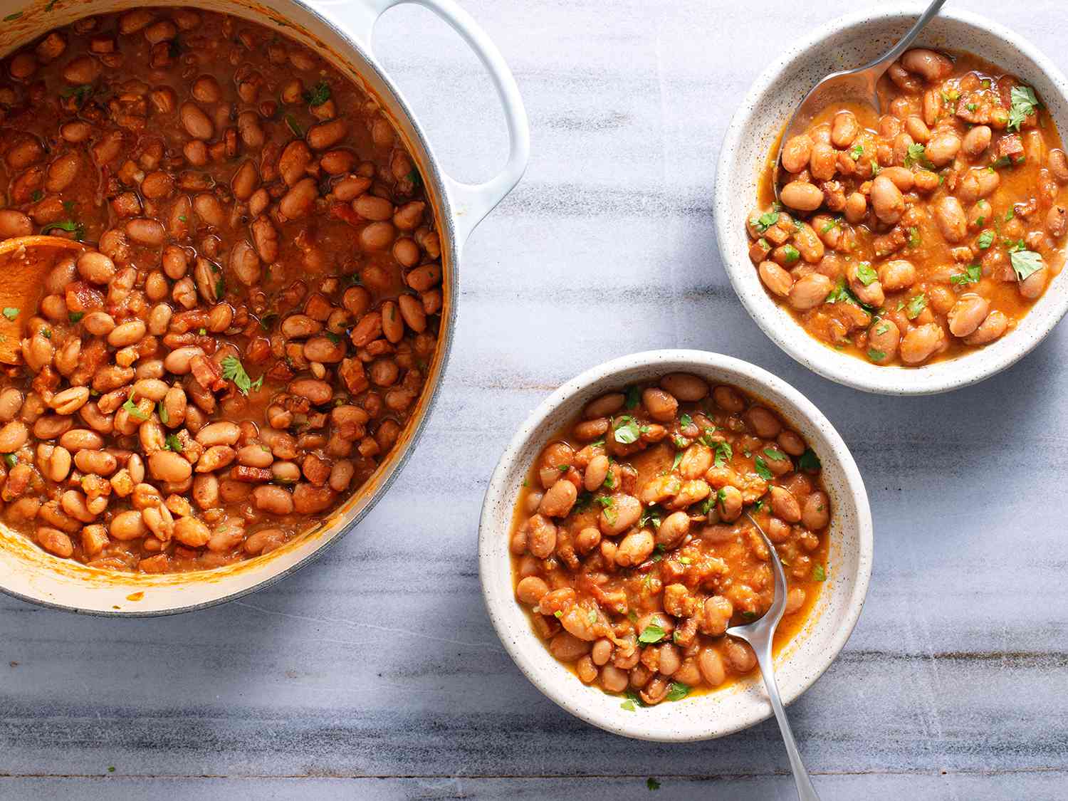A white dutch oven and two white ceramic bowls, all filled with cooked beans, on a white stone background.