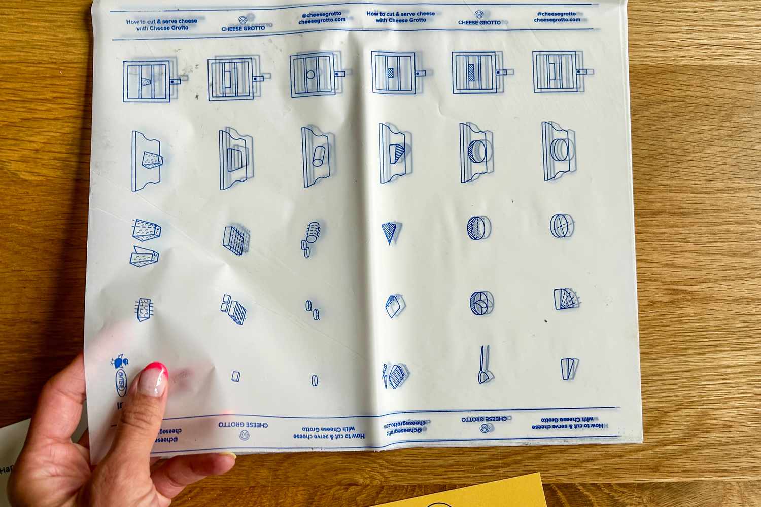 A hand holding a cheese cutting and serving guide, from Cheese Grotto's Monthly Cheese Subscription, displaying visual instructions for slicing various types of cheese