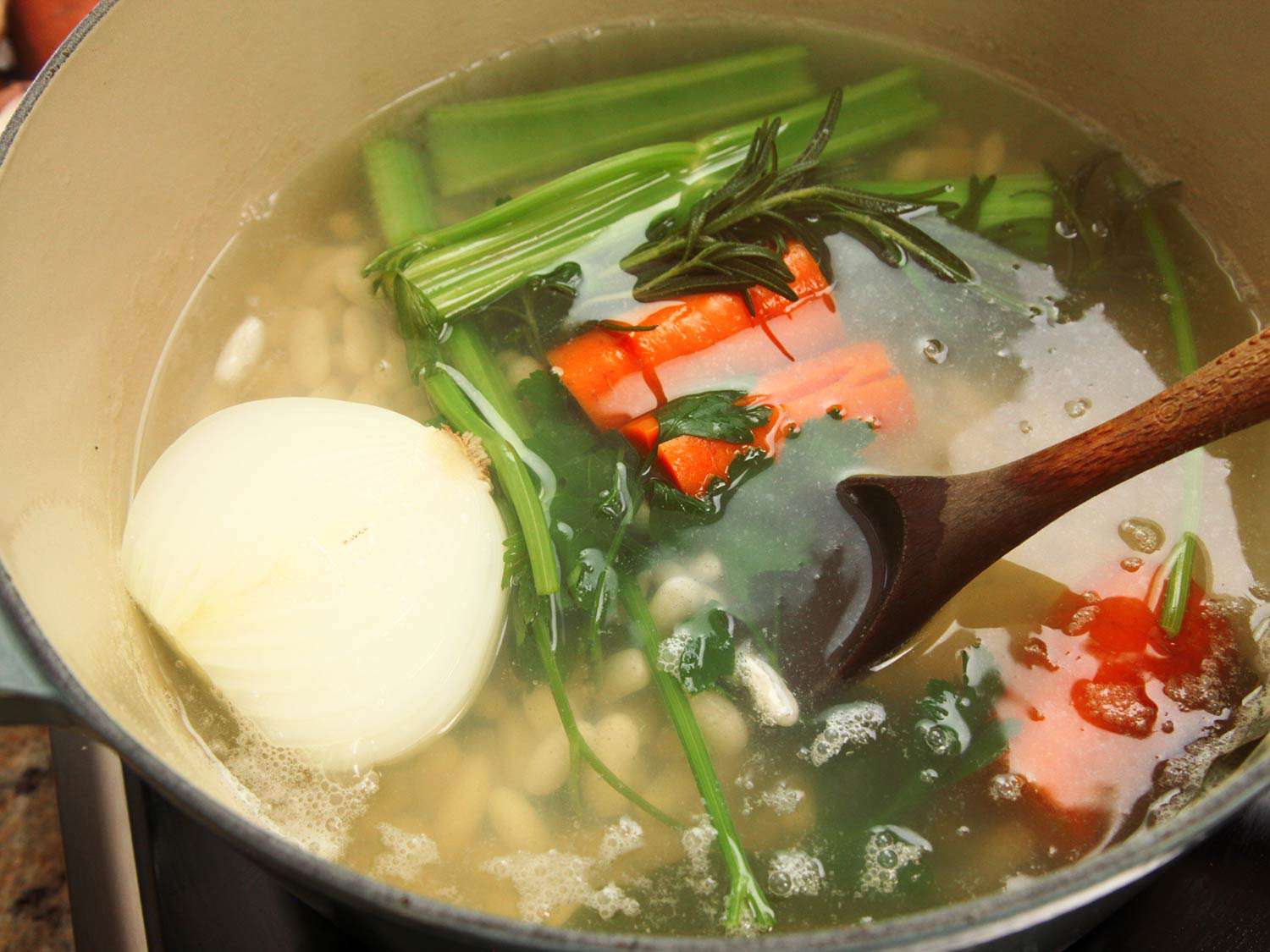 Beans, onion halves, carrot, celery, garlic cloves, rosemary, parsley, and bay leaf simmering in a liquid in a Dutch oven.