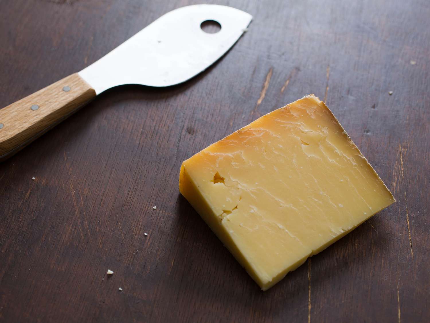 One wedge of hard cheese with a cheese knife next to it. Both are on a dark wood table.