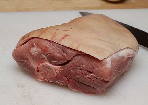 A raw bone-in pork shoulder roast on a white cutting board with a chef's knife. 