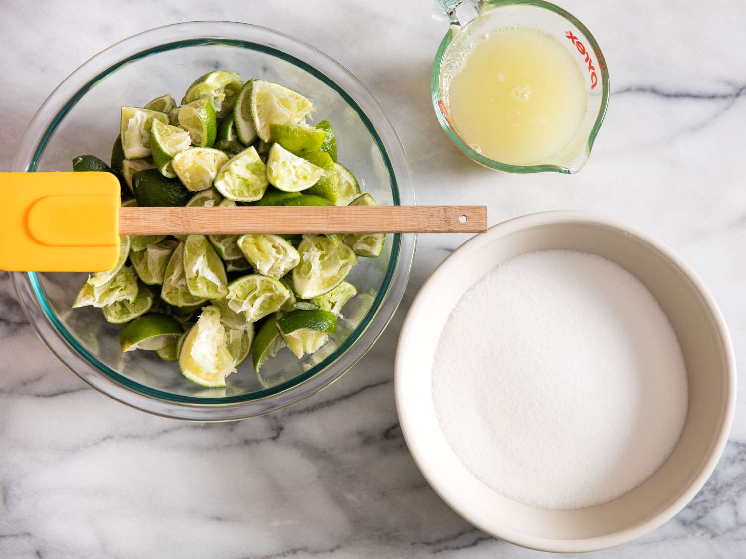 Overhead view of a liquid measuring cup of lime juice, a bowl of spent lime quarters, and a bowl of sugar.