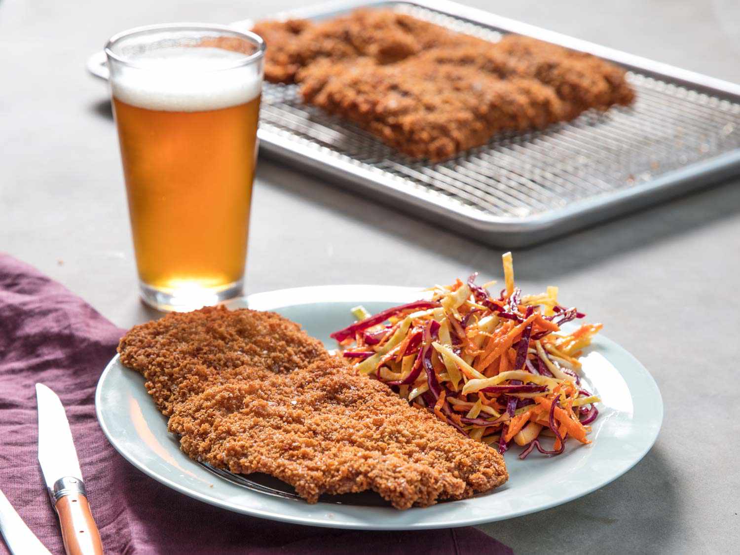 A deep fried breaded pork cutlet on a plate with slaw, with a pint of beer and more pork in the background.