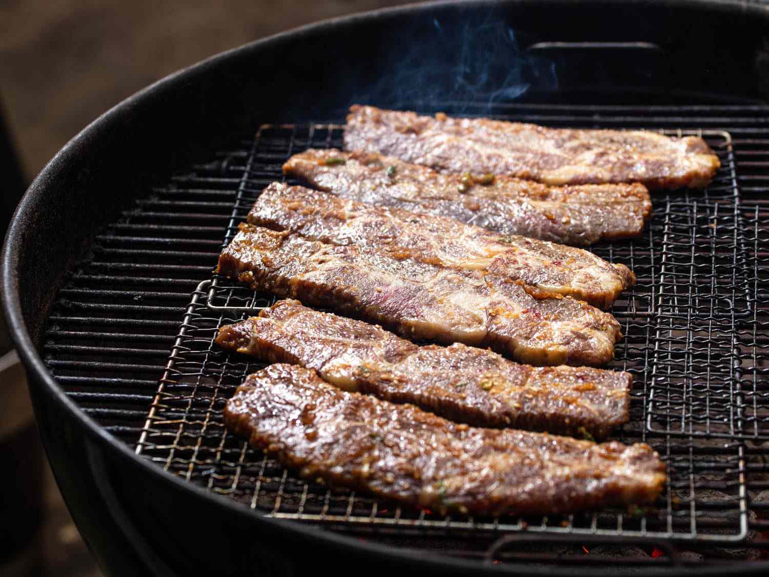 Beef galbi cooking on wire mesh racks set on a charcoal kettle grill.