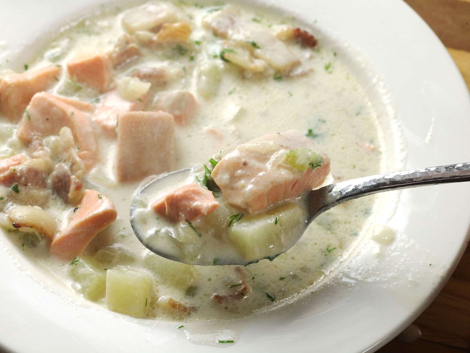 A spoonful of salmon chowder is held up to the camera, inches above a white bowl.