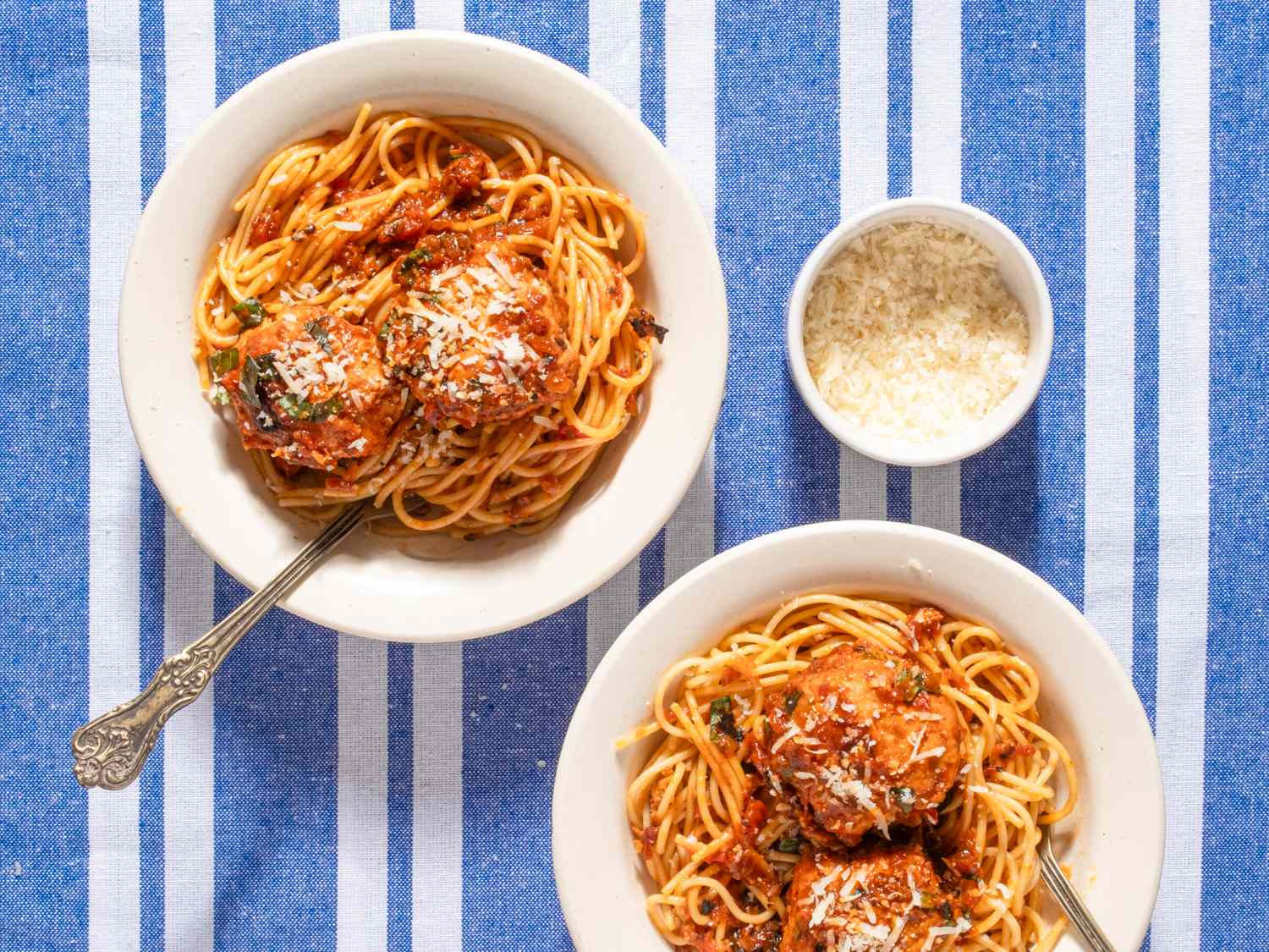 Two bowls of spaghetti with chicken meatballs Parmesan cheese on top and a small bowl of grated Parmesan on a blue striped tablecloth