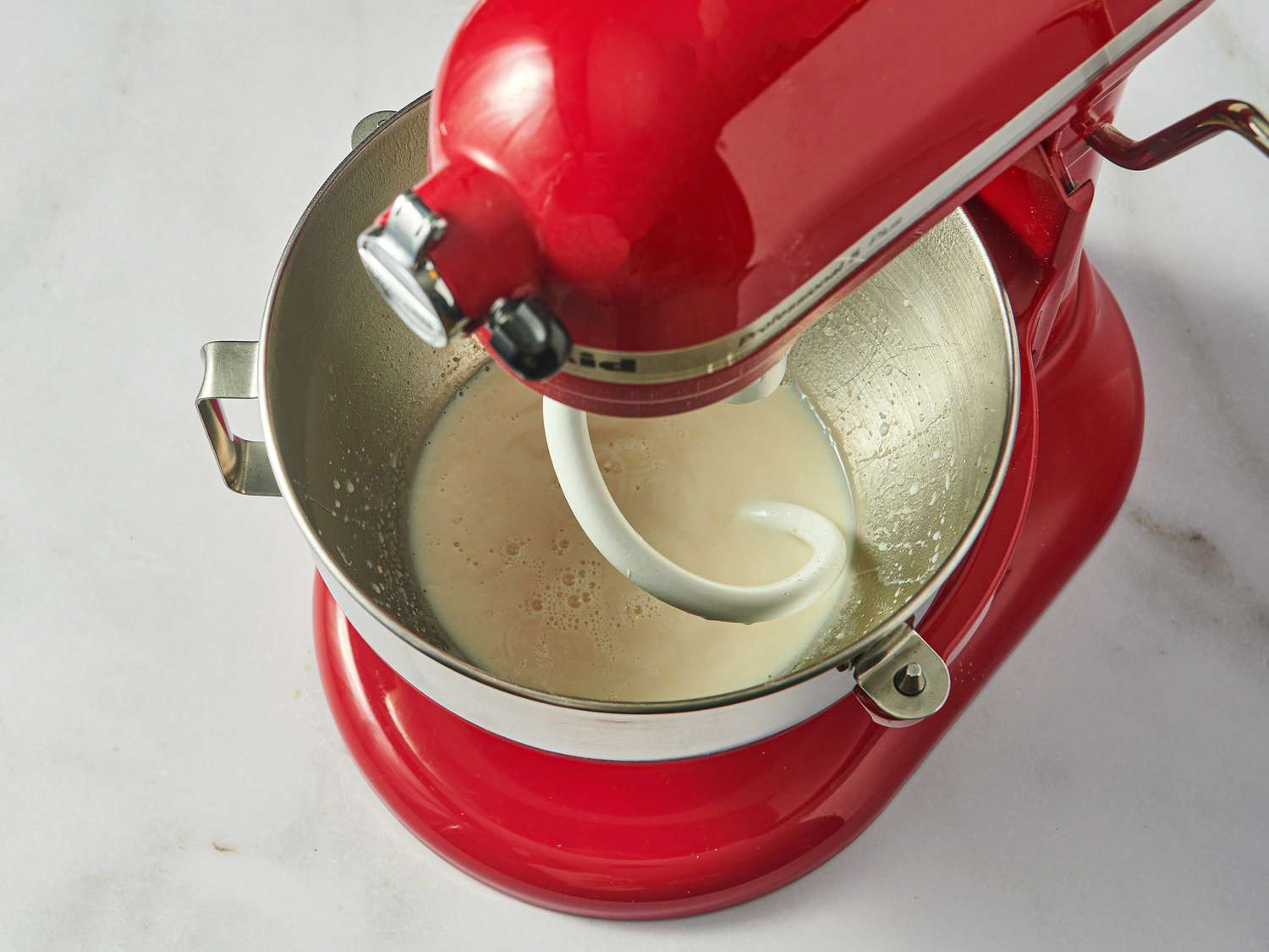 A red stand mixer with a dough hook attachment is mixing ingredients in a metal bowl