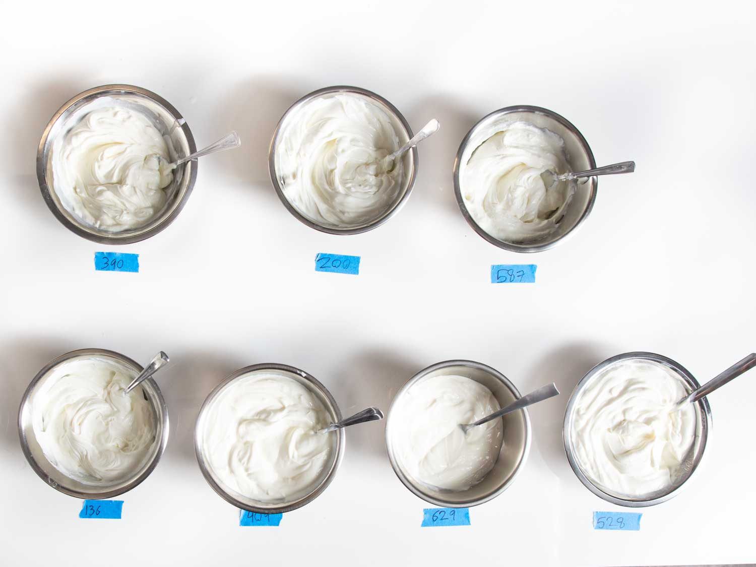 Overhead shot of seven dishes of Greek yogurt to be tasted, each with a spoon stuck inside, and a numbered blue label placed beneath each one.