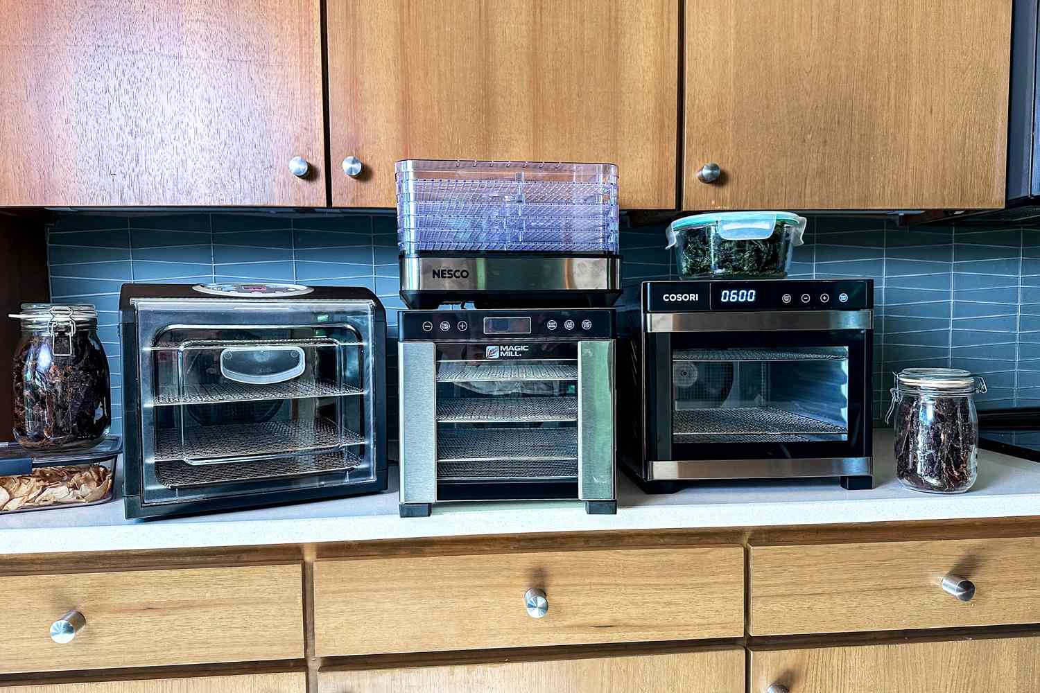 Four food dehydrators on a kitchen counter with jars of beef jerky and kale chips next to them