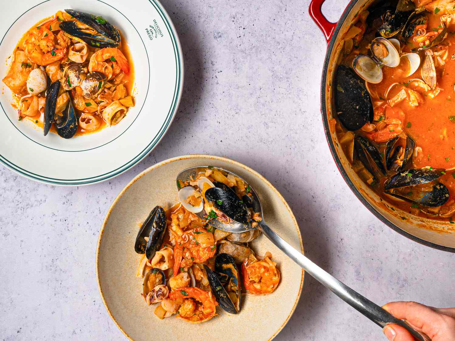 A hand serving seafood stew from a pot into a bowl featuring mussels shrimp and clams on a table with plated portions of the dish