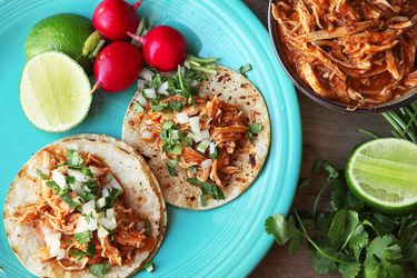 Two tacos topped with chicken tinga, diced onion, and cilantro. There are radishes and cut limes on the plate and a dish of chicken tinga nearby. 