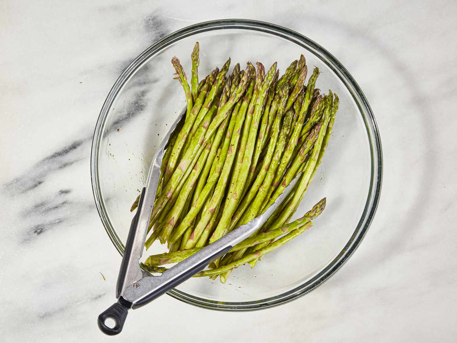 Asparagus, oil, and seasonings are tossed in a tempered glass bowl with tongs.