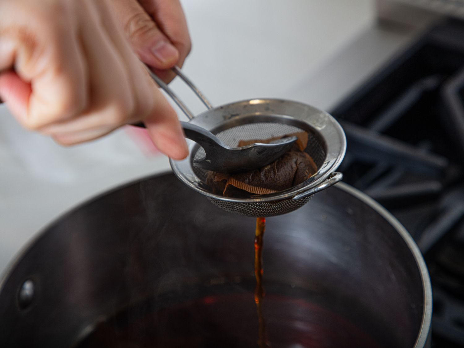 Tea being strained with a metal strainer and spoon over a cooking pot