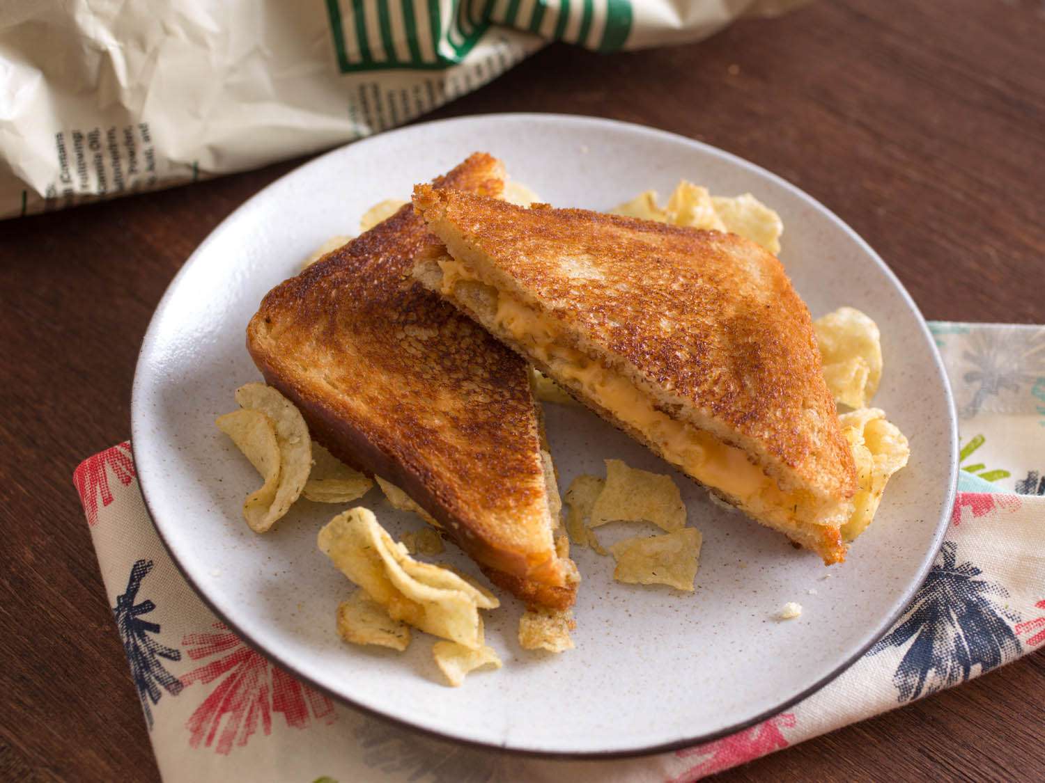 A grilled cheese sandwich cut in half on a plate with the two halves slightly stacked. The plate is scattered with potato chips and is sitting on a wooden table, with a cloth napkin under the plate.