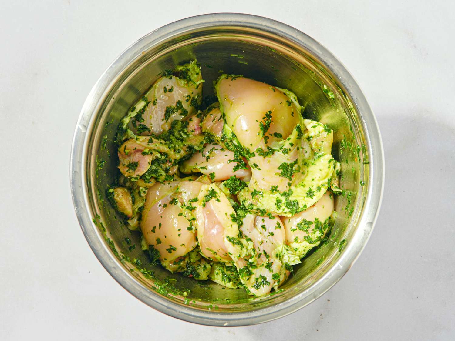 Raw chicken thighs mixed with chimichurri sauce in a metal bowl