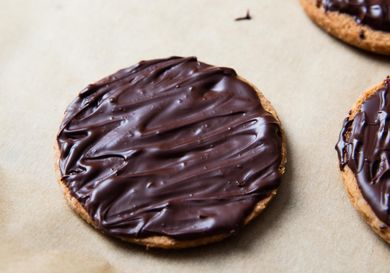 A vegan chocolate-covered digestive biscuit resting on parchment paper. There are two other biscuits on the right periphery of the image.