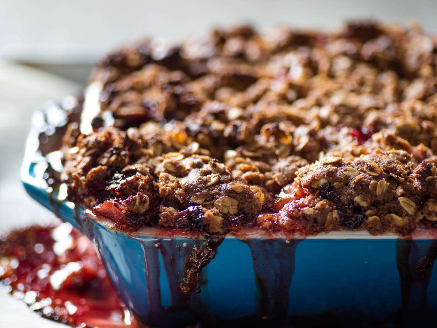 Close-up of a blue ceramic baking dish of strawberry-rhubarb crisp, with fruit filling spilling over the side