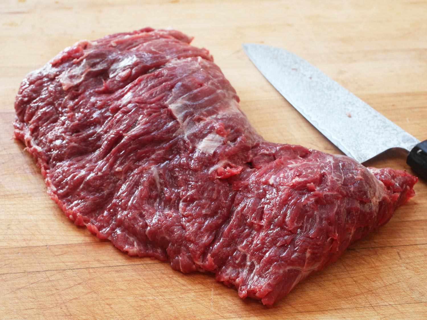 Raw flap meat steak on a wooden cutting board with a knife in the background.
