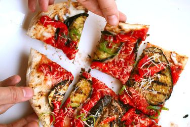 A grilled pizza, sliced into pieces and being taken by several hands on a white background.