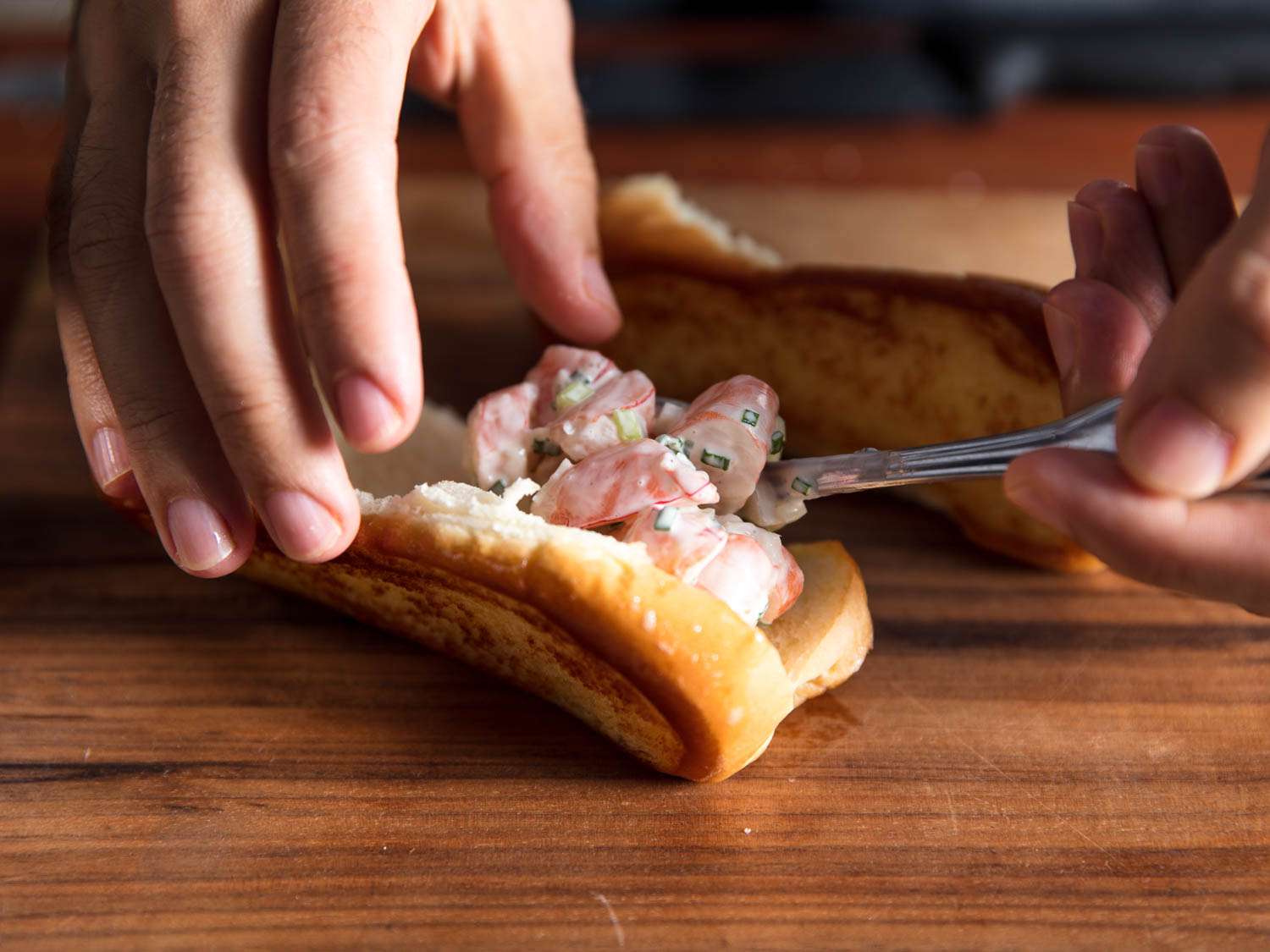 Spooning shrimp mixed with a mayo-based dressing into buttered, toasted buns for shrimp rolls.