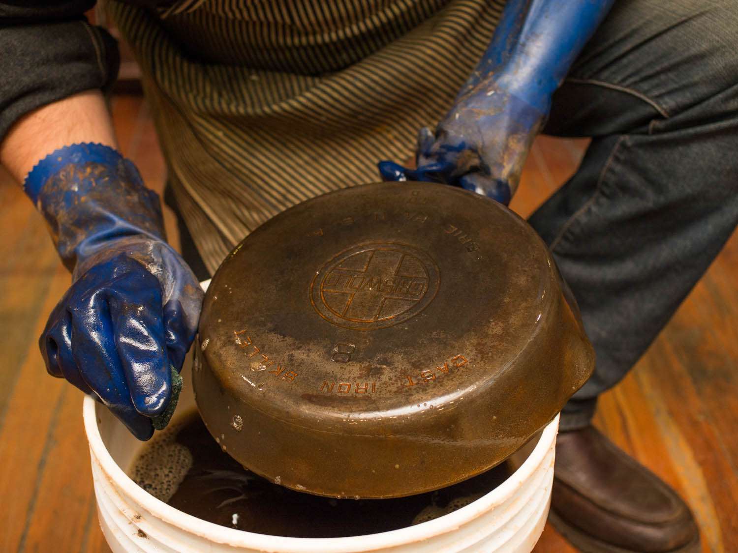 A pair of gloved hands scrubbing an old cast iron over a white bucket.