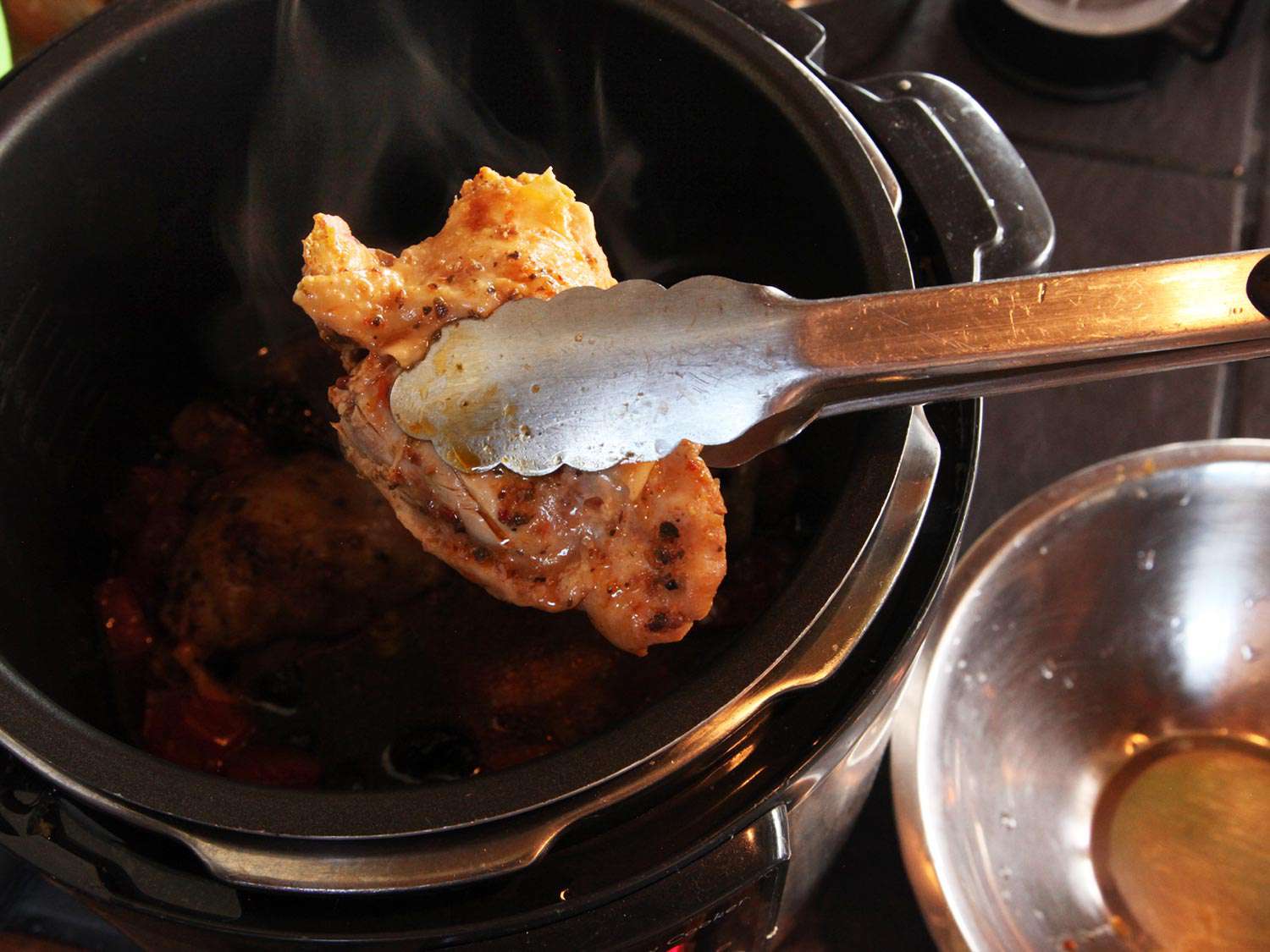 A braised chicken thigh is removed from an electric pressure cooker with tongs.