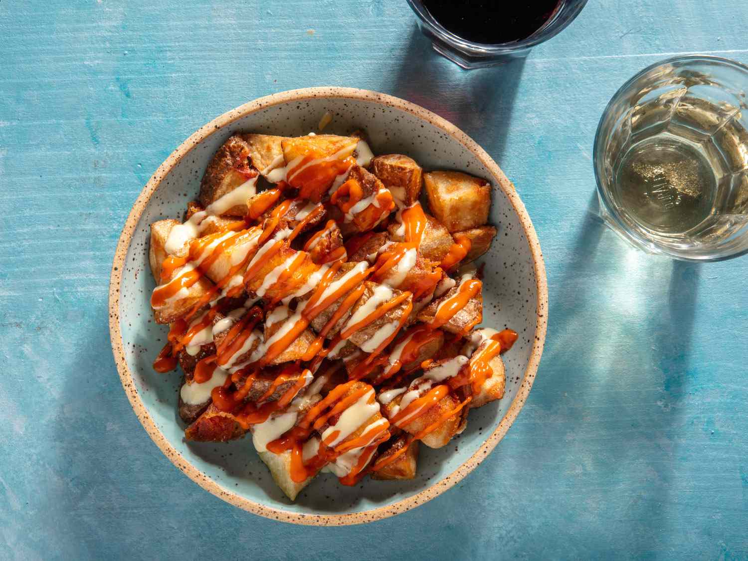 Overhead of a small serving bowl of patatas bravas drizzled with salsa brava and allioli, with glasses of red and white wine on the side.