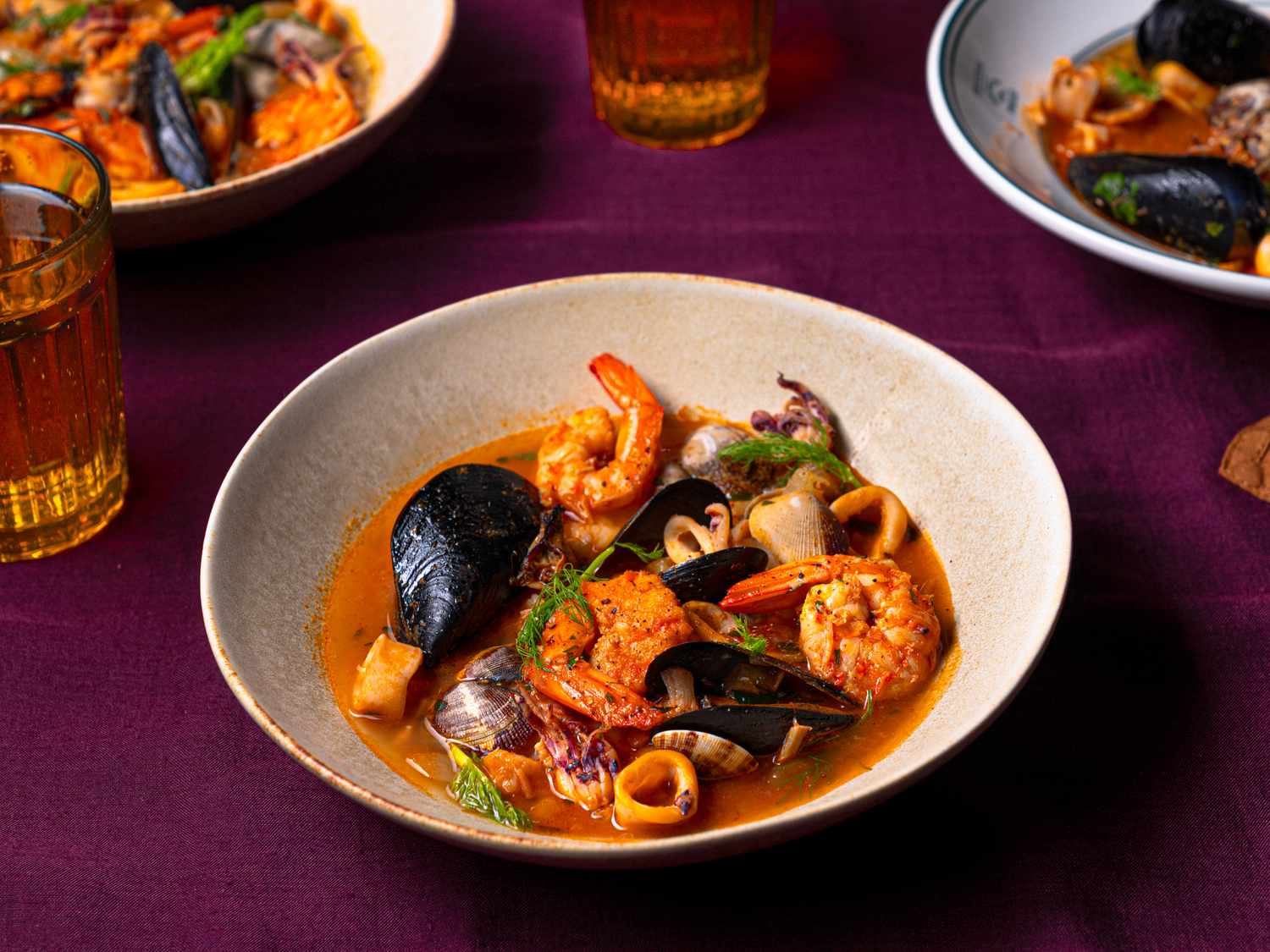 A seafood dish in a white bowl with shrimp mussels and other seafood placed on a dark tablecloth near a glass of ambercolored liquid