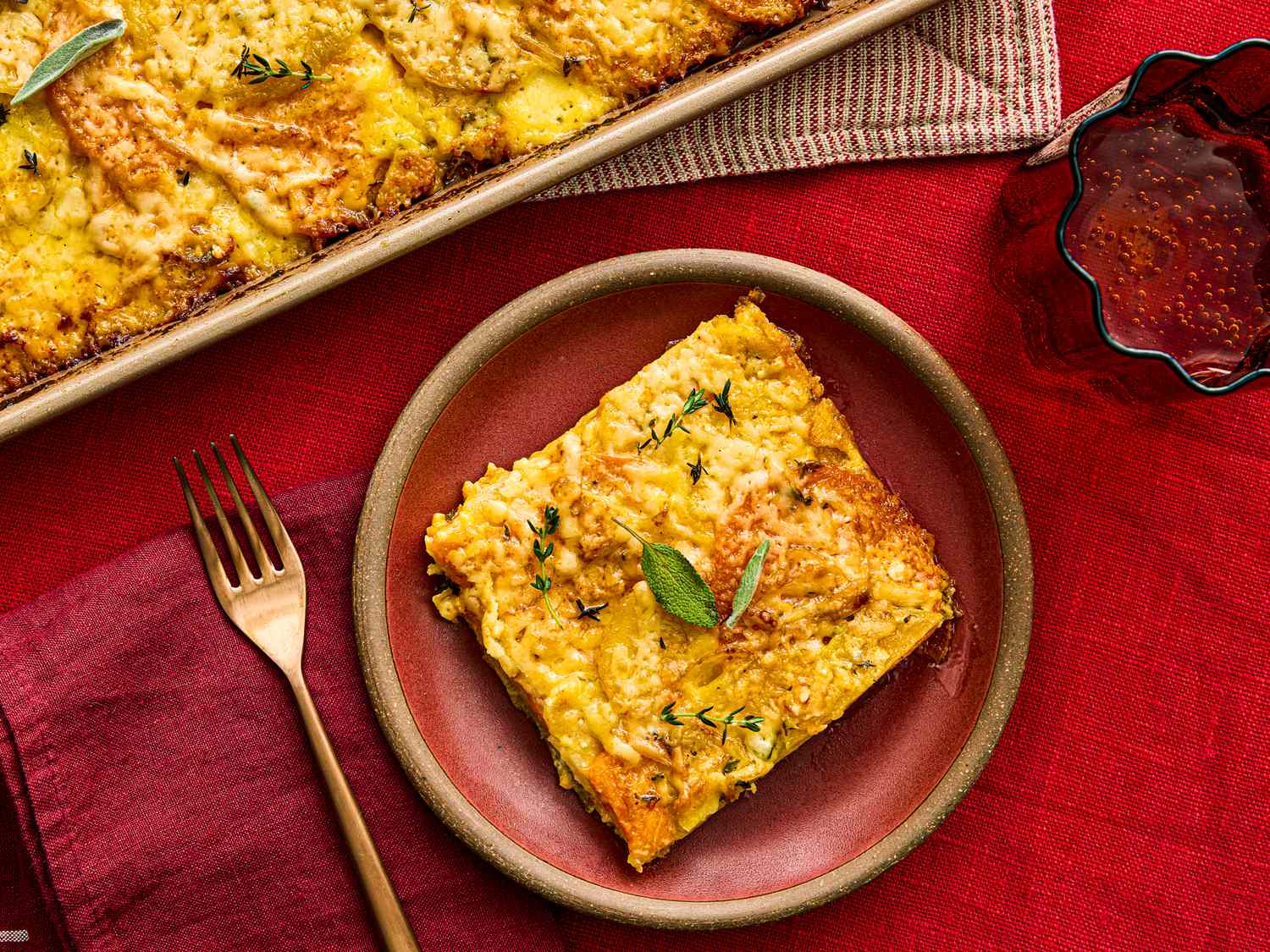 A serving of root vegetable gratin on a plate with a fork garnished with herbs and set on a red background