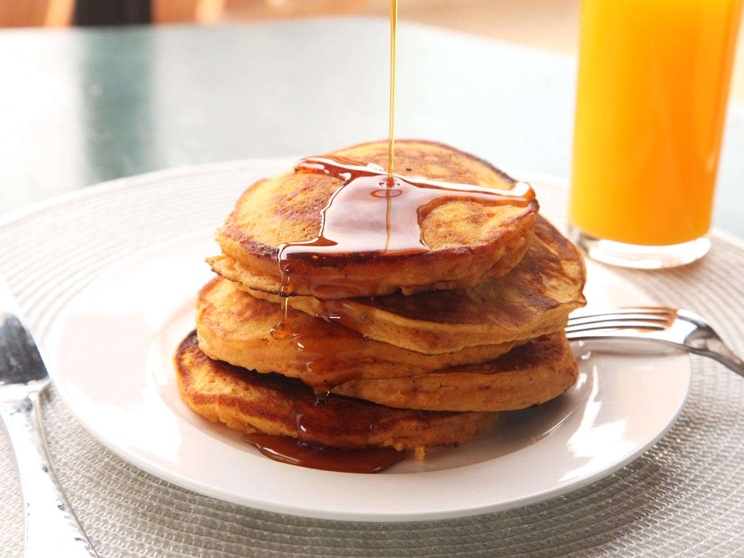 More maple syrup being poured over a stack of sweet potato pancakes on a white plate with a fork next to a glass of orange juice.