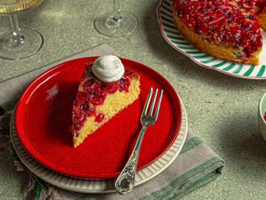 A slice of cranberry upsidedown cake on a red plate with a dollop of cream fork placed alongside the slice whole cake in background