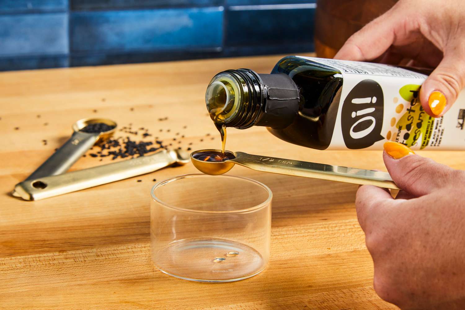 Pouring oil into a measuring spoon with a wooden surface and black sesame seeds in the background