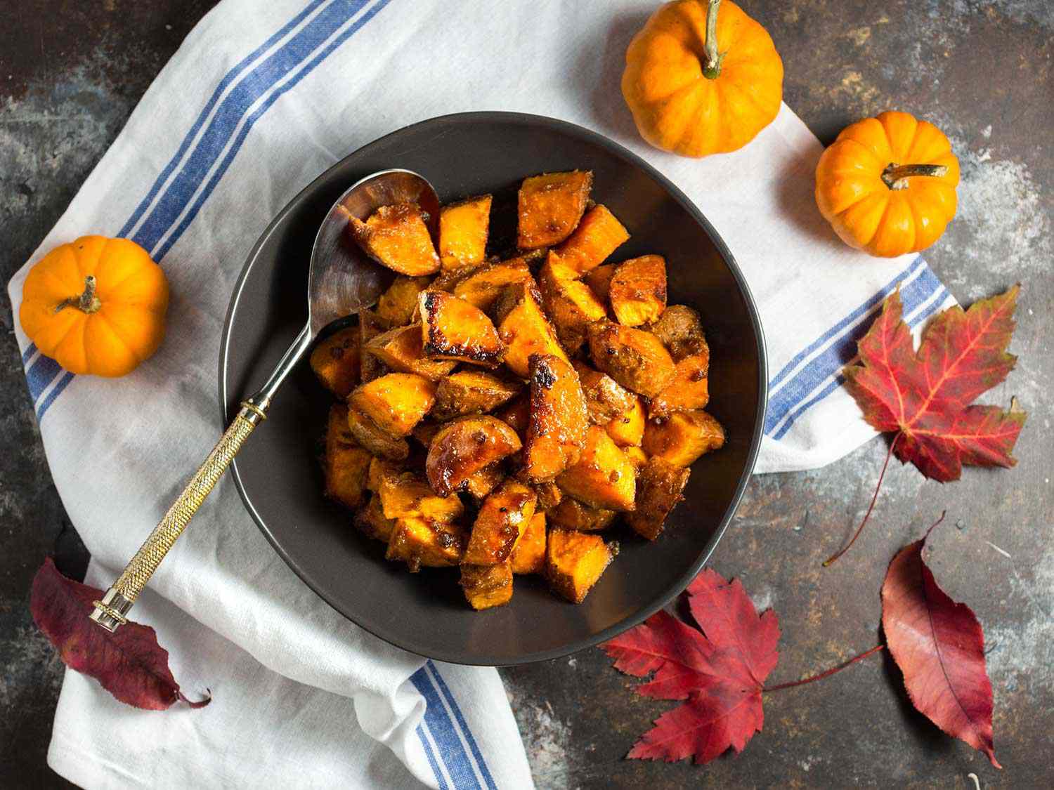 An overhead view of a serving bowl full of roasted sweet potatoes with miso butter and maple. Maple leaves are strewn on the countertop, and small decorative squash are perched nearby.