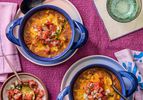 Overhead of 2 blue bowls with taco soup, on with a spoon in it. A small dish with pico de gallo on it. Pink napkins and sparkling water and a magenta colored textile surface. 