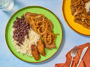Green Plate of Pallomilla with rice beans and plantains. Red napkin with untensils, and large plate of pallomilla to the side 