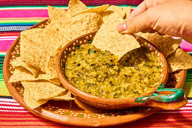 Mexican clay bowls with tortilla chips and charred salsa verde on a colorful traditional Mexican textile, with a hand dipping a chip into the salsa 