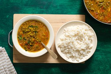 Bowl of Moong dal in a bowl, with a bowl of white rice next to it. Bowls are on a wooden board on top of green textured surface, and larger bowl of Dal is in the side of the frame. 
