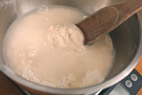 Wooden spoon beginning to mix flour into water in silver mixing bowl.