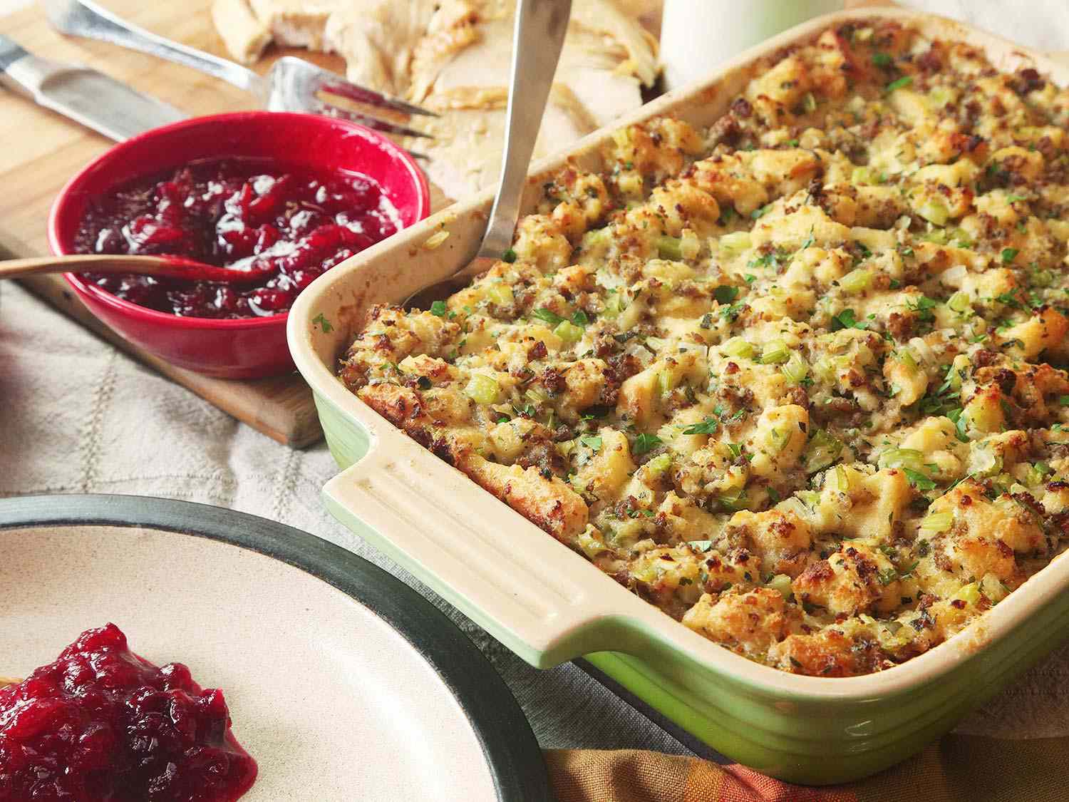  A baking dish of Thanksgiving stuffing with a spoon sticking out of it, next to turkey and a bowl of cranberry sauce.