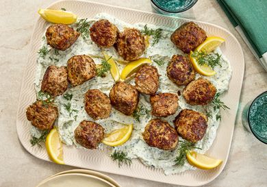 meatballs on top of yogurt with fresh herbs and lemon wedges on a platter. Side of sparkling water and green napkin on the side 
