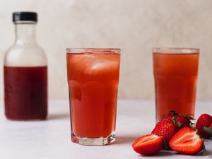 Two glasses of berry shrub. On the right hand side of the image are some strawberries, both whole and sliced in half, and on the left side of the image, in the background, is a small glass bottle holding more shrub.