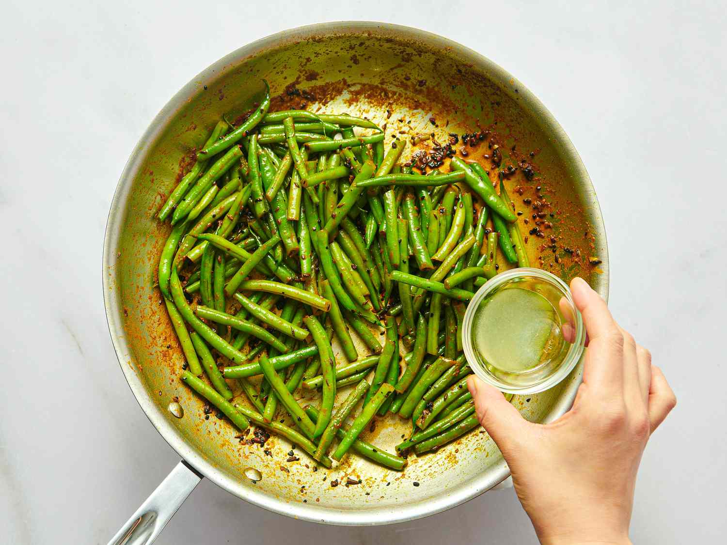 Cooking green beans in a pan with a liquid being poured from a bowl