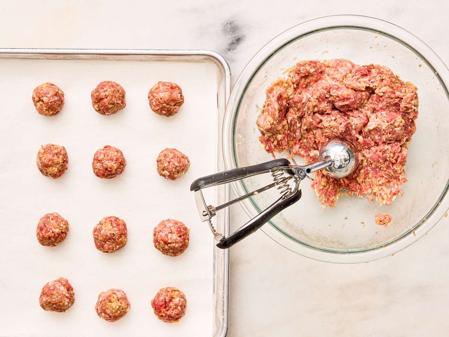 Prepared raw meatballs on a tray and a bowl of meat mixture with a scoop