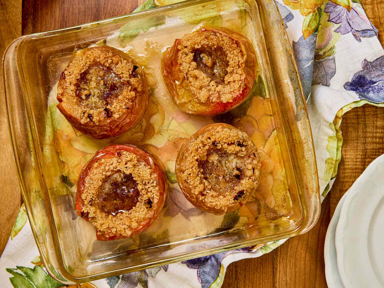 Four stuffed baked apples in a clear rectangular baking dish sitting on a colorful floral patterned cloth on a wooden surface