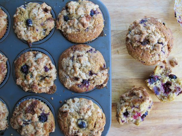 An overhead view of six berry muffins with a streusel topping in a pan on a wooden table, with one whole muffin and one split in half next to it.