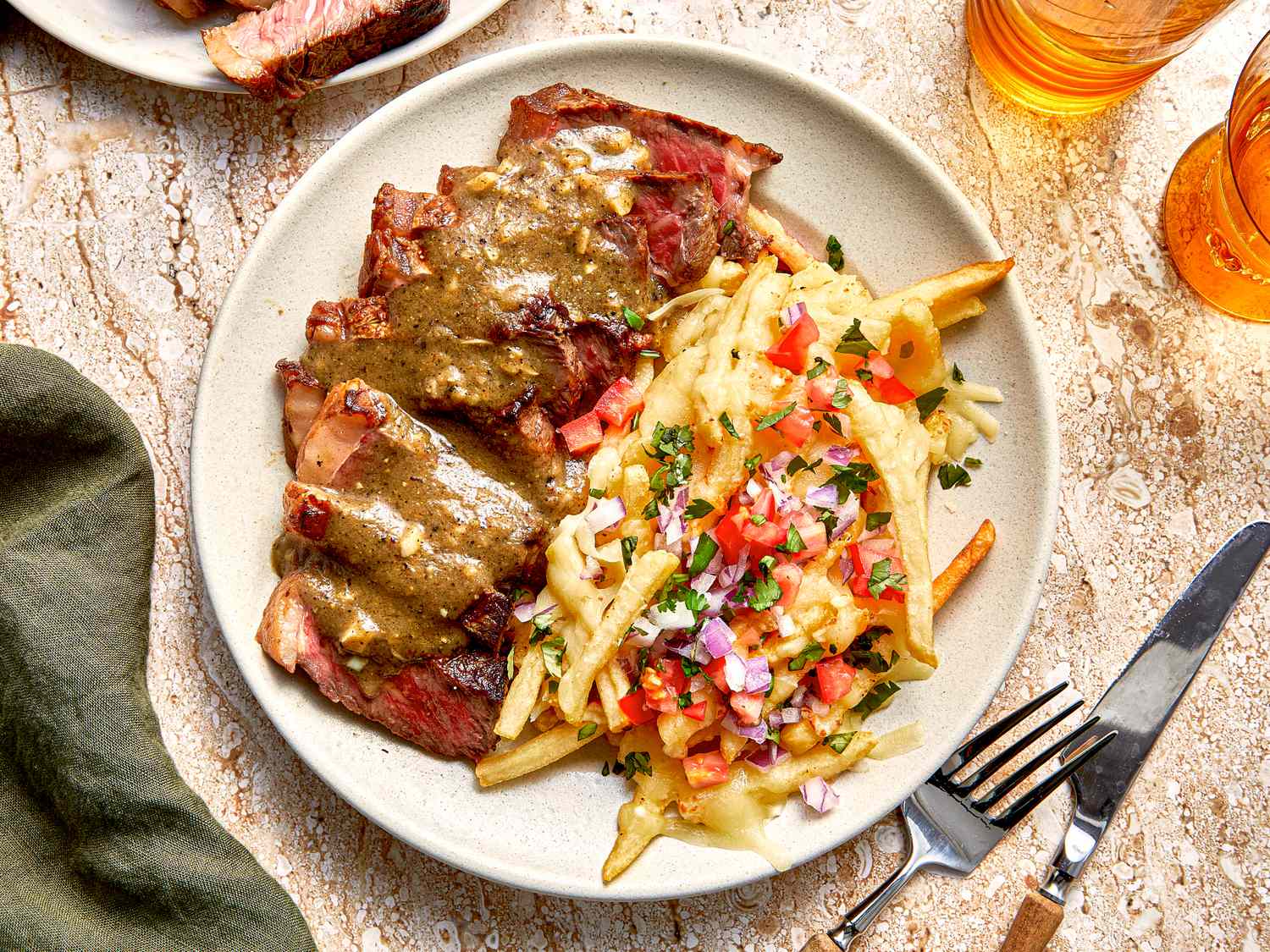 A plate with sliced steak covered in sauce and loaded fries topped with cheese and diced toppings utensils on the side