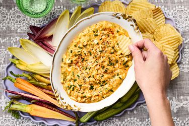 A hand dipping a potato chip into deviled egg dip, surrounded by vegetables on a tray