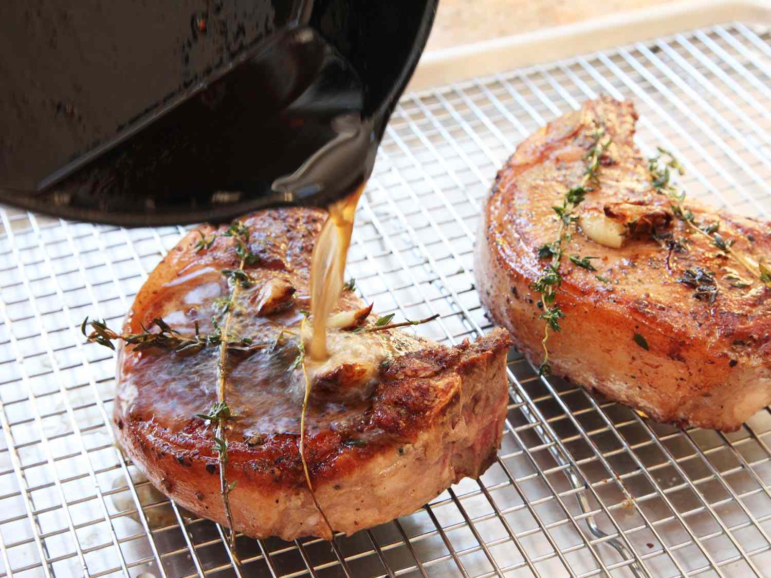 Two cooked pork chops resting on a cooling rack as their juices are poured over them from a cast iron skillet.