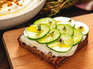Openfaced sandwich with sliced cucumber on bread with a white spread topped with herbs served on a wooden surface